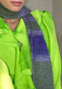 Blusa verde com babados, acompanhada de um lenço listrado em cinza e roxo. O lenço apresenta uma textura suave e fofinha, com uma transição de cores em degradê.