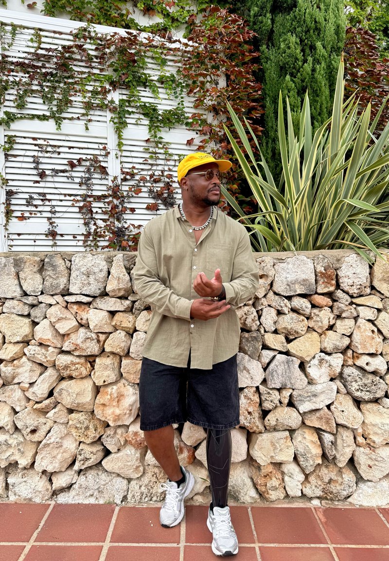 Man with a prosthetic leg wearing black shorts, an olive shirt, a yellow cap, and sunglasses stands beside a stone wall adorned with green and variegated plants.