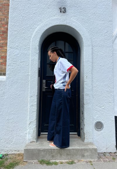 Pantalones de mezclilla de pierna ancha en azul oscuro, con un ajuste holgado. Combinados con una camiseta de jersey blanca con acentos rojos y azules, de pie junto a una pared texturizada.