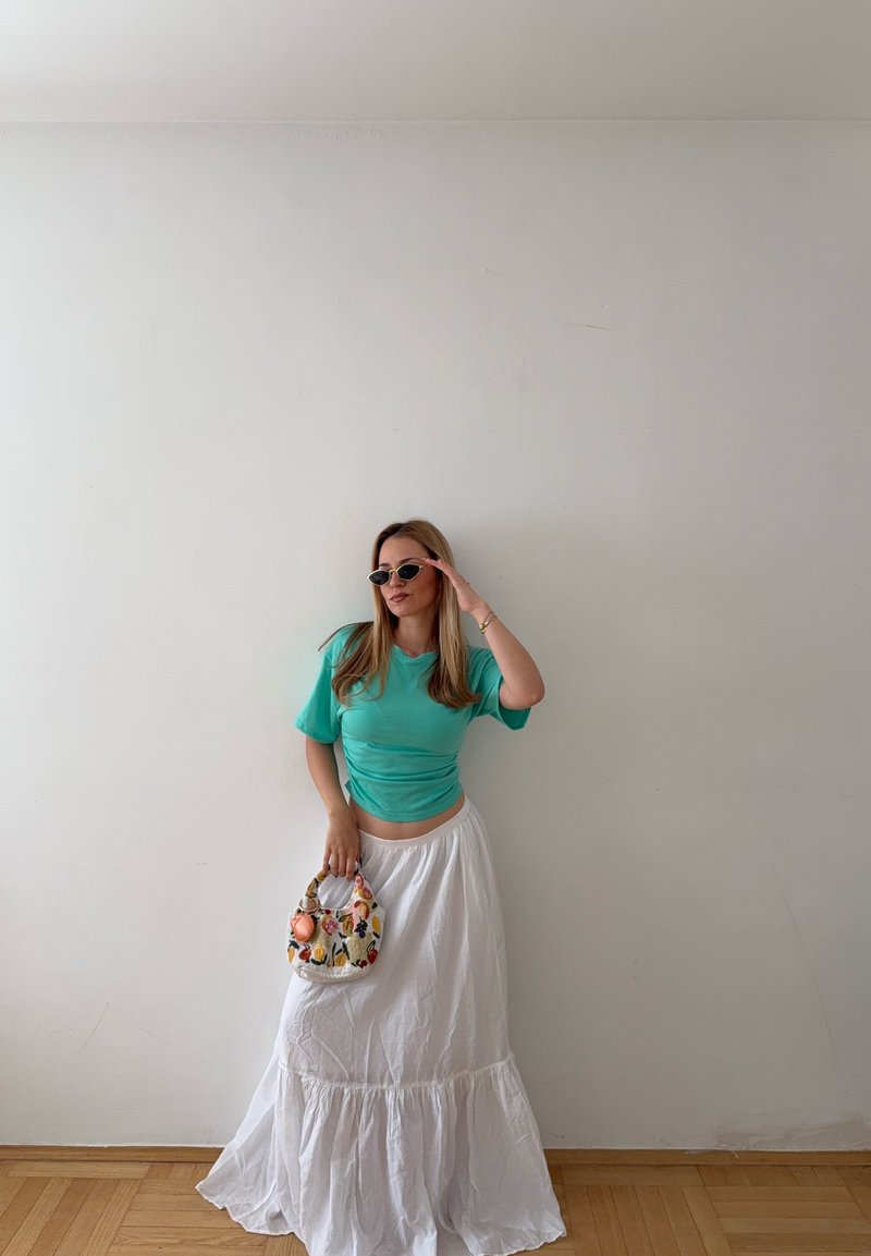 Femme portant un top bleu sarcelle et une longue jupe blanche tient un sac à main fleuri et ajuste ses lunettes de soleil devant un mur uni.