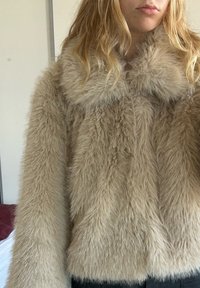 Blond person iført en beige, blød imiteret pelsjakke, vist fra skuldre til talje, stående indendørs mod en ensfarvet væg.