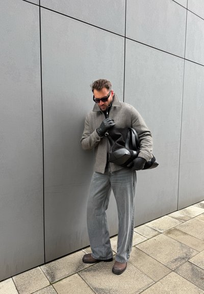 Hombre con gafas de sol, chaqueta de cuadros, pantalones grises, guantes negros y botas, sosteniendo una bolsa negra grande frente a una pared gris de azulejos.