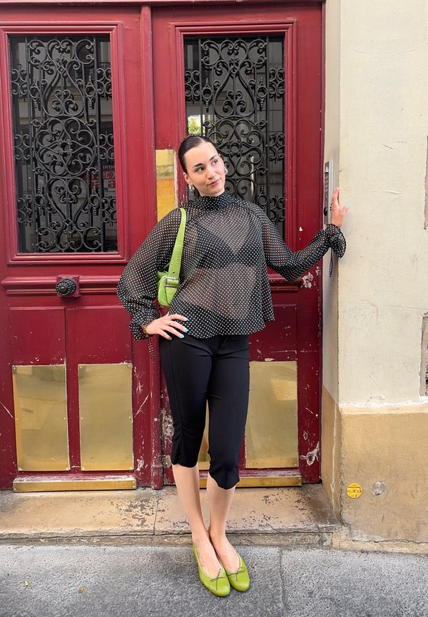 Femme en blouse noire transparente à pois, pantalon noir, chaussures et sac verts, appuyée contre une porte rouge ornée de panneaux en fer forgé.