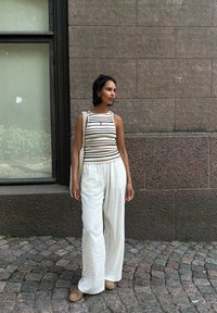 Top sin mangas a rayas en crema y negro, combinado con pantalones anchos blancos y sueltos. Bolso de mimbre y zapatos beige tipo slip-on. Fondo de pared de piedra.