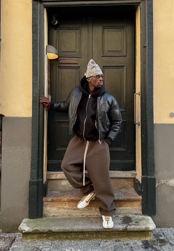 Black leather bomber jacket over a black hoodie, beige sweatpants with white drawstring, gray patterned beanie, standing by a dark green door.