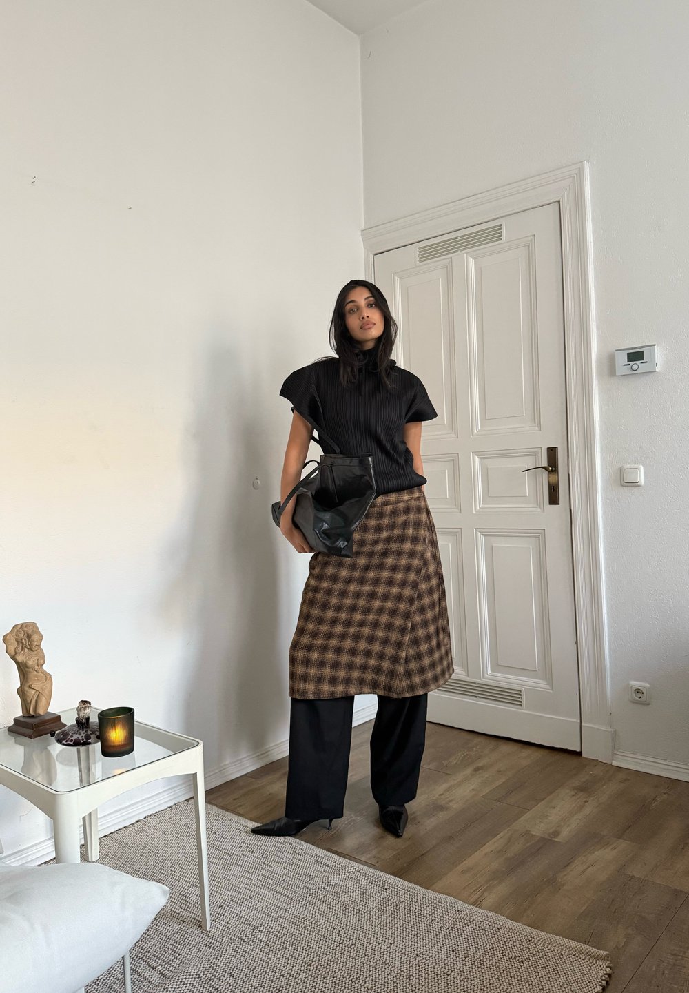 Fekete bordázott felső, barna kockás szoknya, fekete bőszárú nadrág, fekete táska, hegyes sarkú cipő. Világos fa padló és egyszerű fehér falak a háttérben.