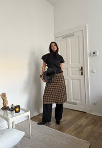 Fekete bordázott felső, barna kockás szoknya, fekete bőszárú nadrág, fekete táska, hegyes sarkú cipő. Világos fa padló és egyszerű fehér falak a háttérben.