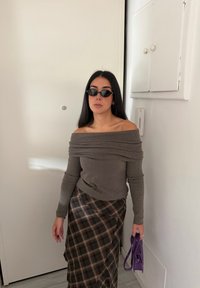 Szary sweter odkrywający ramiona z opadającym dekoltem, zestawiony z brązową spódnicą w kratę, zapinaną na zakładkę. Trzyma małą fioletową torebkę.