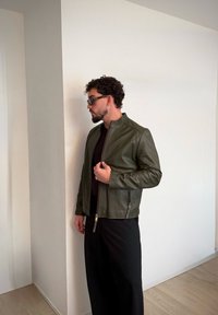 Olívazöld bőrdzseki álló gallérral, cipzáras zsebekkel, sima textúrával és karcs�úsított szabással, fekete nadrággal párosítva.