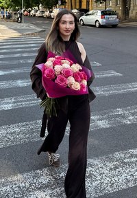 Bukiet różowych i kremowych róż owinięty w fioletowy papier, trzymany przez osobę w ciemnym ubraniu, przechodzącą przez ulicę na pasach.