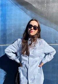 Camicia azzurro chiaro con accenti in tessuto trasparente che ricordano fiocchi, indossata con occhiali da sole neri oversize su uno sfondo blu.