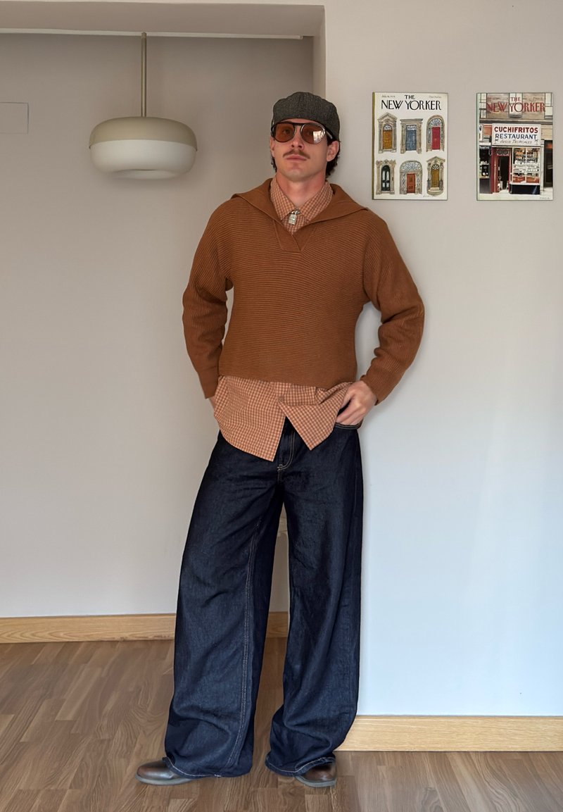 Homme portant un pull marron par-dessus une chemise à carreaux, un pantalon large foncé, des lunettes de soleil et une casquette plate, debout à l'intérieur contre un mur avec des affiches encadrées.