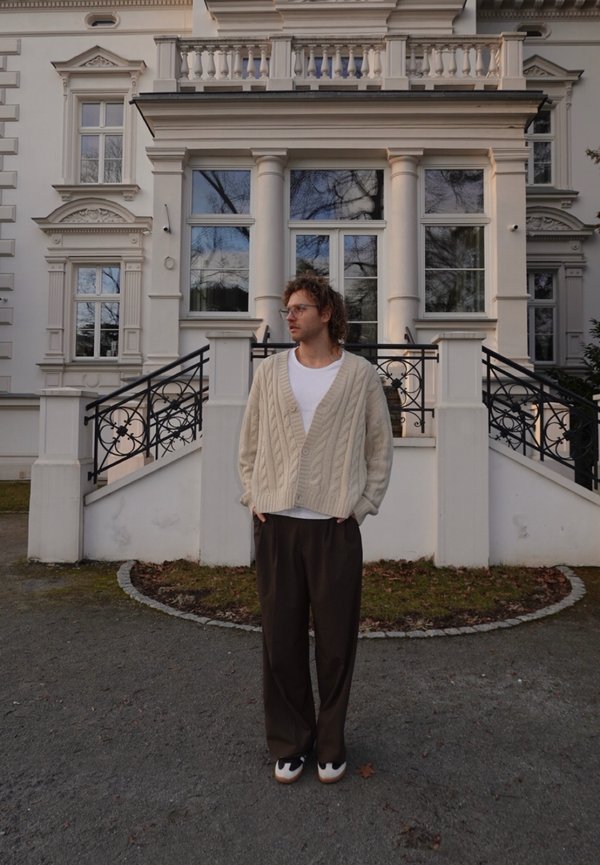 Homme portant un cardigan beige en maille torsadée, une chemise blanche et un pantalon foncé, debout devant un bâtiment classique blanc avec des colonnes et des rampes en fer.