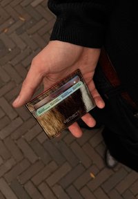 Card holder maro și crem, realizat din piele texturată, cu trei sloturi pentru carduri, logo embossat, ținut într-o mână pe un fundal din cărămidă.