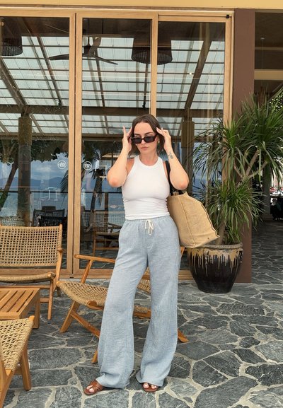 Top de tirantes blanco con estrías, pantalones de pierna ancha a rayas azul claro con cordón, sandalias marrones, bolso grande beige, en un entorno tropical con vegetación.