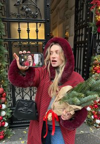 Manteau à carreaux rouge et noir avec une capuche doublée de fourrure ; tenant un petit bouquet de conifères enveloppé dans du papier kraft et un sac à main noir.
