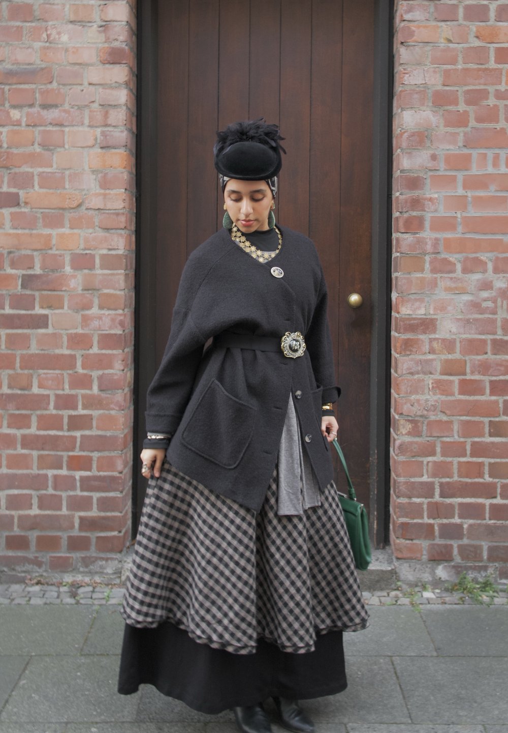 Femme portant un chapeau noir, un grand cardigan foncé avec des broches décoratives, une jupe à carreaux, tenant un sac à main vert, debout devant une porte en bois et un mur de briques.