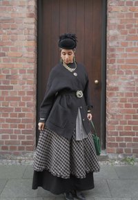 Femme portant un chapeau noir, un grand cardigan foncé avec des broches décoratives, une jupe à carreaux, tenant un sac à main vert, debout devant une porte en bois et un mur de briques.