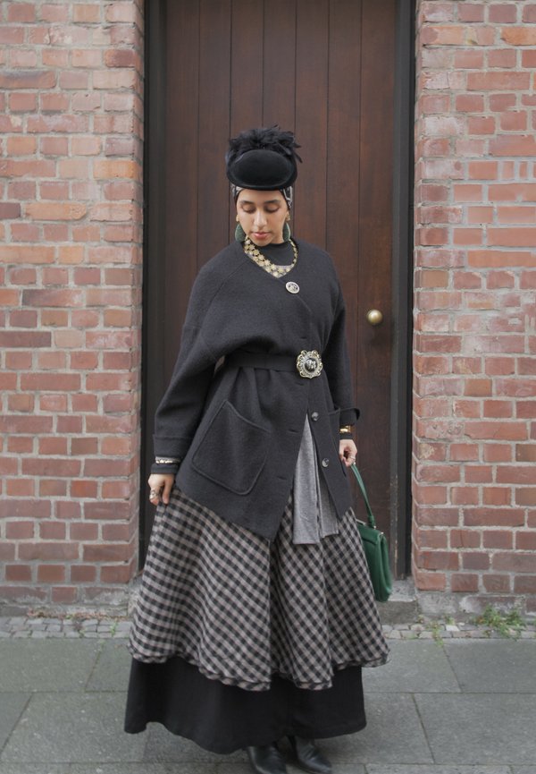 Femme portant un chapeau noir, un cardigan sombre surdimensionné orné de broches décoratives, une jupe à carreaux, tenant un sac à main vert, debout devant une porte en bois et un mur en briques.