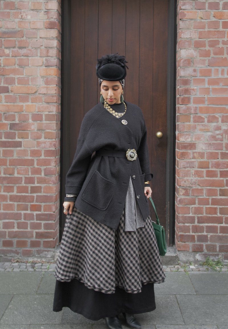 Femme portant un chapeau noir, un grand cardigan foncé avec des broches décoratives, une jupe à carreaux, tenant un sac à main vert, debout devant une porte en bois et un mur de briques.