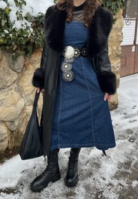 Personne portant un manteau en cuir noir avec col et poignets en fourrure, robe en denim bleu, ceinture large décorative, bottes de combat noires, tenant un sac noir, debout sur la neige.