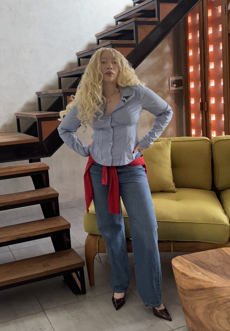 Γυναίκα με μακριά ξανθά σγουρά μαλλιά στέκεται με τα χέρια στη μέση κοντά σε ξύλινη σκάλα και πράσινο καναπέ σε μοντέρνο σαλόνι.