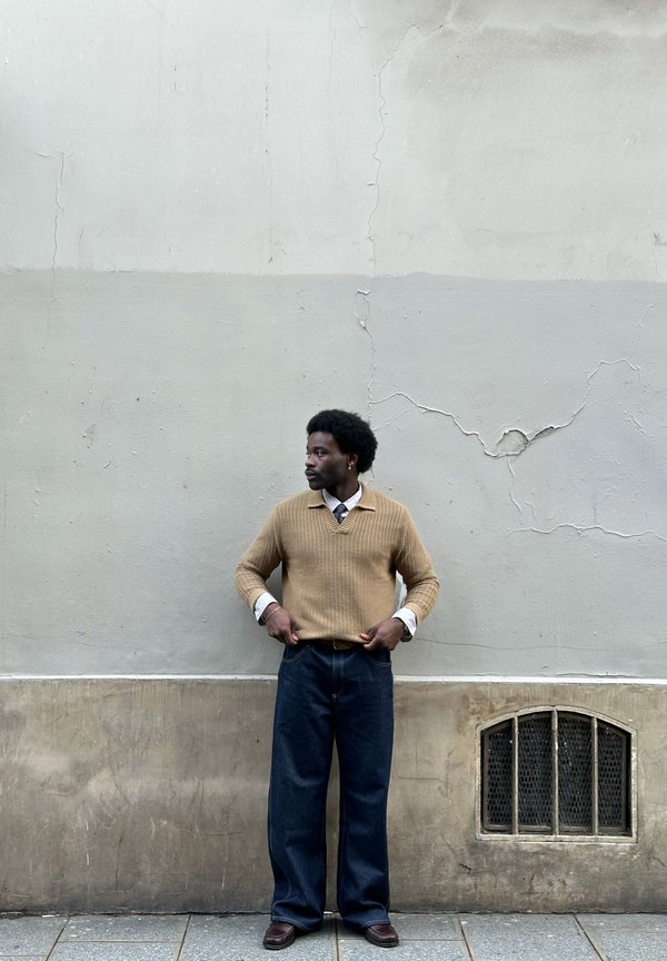Muž v béžovém svetru, bílé košili, tmavě modrých kalhotách a hnědých botách stojící opřený o popraskanou béžovou zeď vedle malého větraného okénka s mříží.