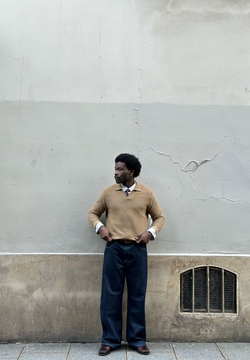 Homme en pull beige, chemise blanche, pantalon bleu marine et chaussures marron, debout contre un mur beige fissuré avec une petite ventilation grillagée à proximité.