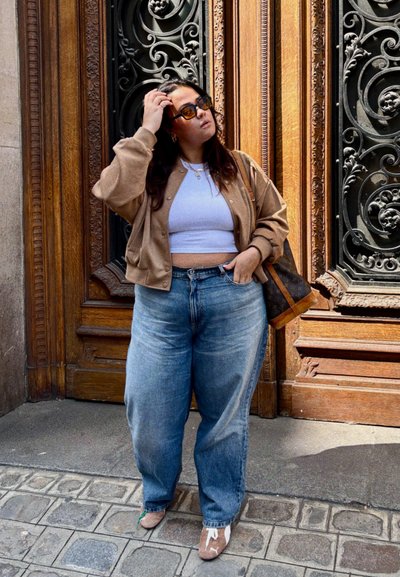 Cárdigan beige sobre un top corto blanco, jeans sueltos azules y zapatos planos estampados; llevando un bolso marrón. Fondo de madera con detalles ornamentales.