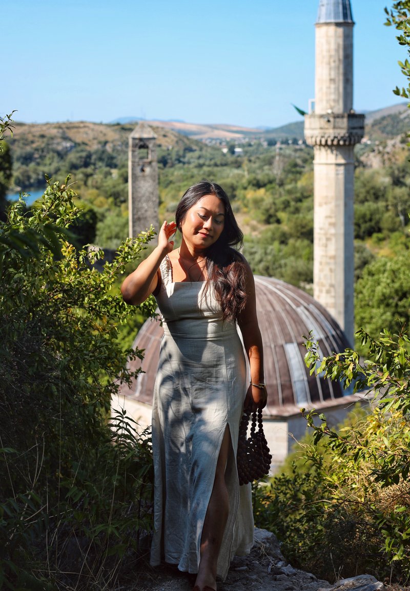 Μάξι φόρεμα σε ανοιχτό μπεζ ύφασμα με πλαϊνό σκίσιμο και λεπτές τιράντες. Μια υφή, σκούρα τσάντα από υφαντό υλικό κρατιέται με το ένα χέρι. Σκηνικό φόντο με πέτρινες κατασκευές.