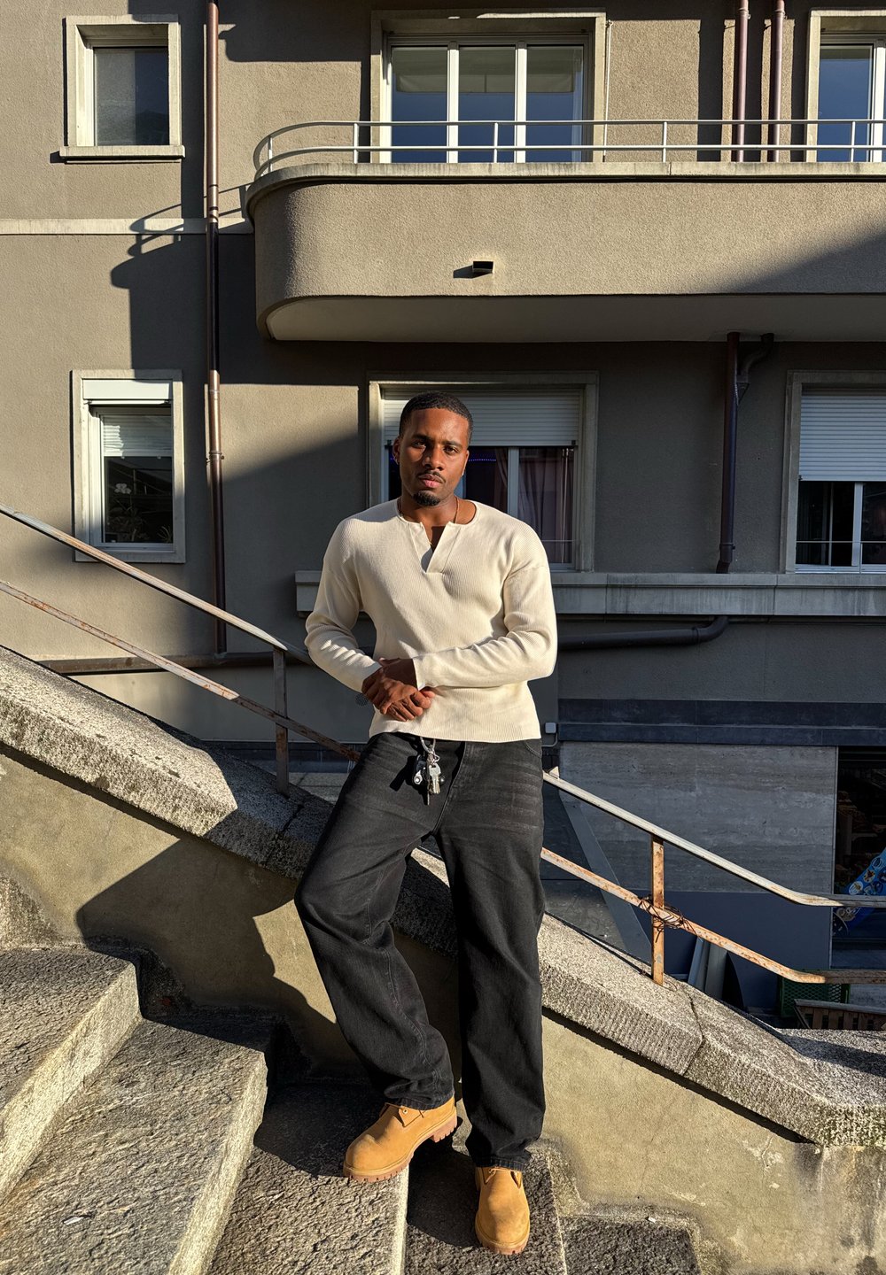 Camisa blanca de manga larga, pantalones negros y botas beige; de pie en unos escalones de piedra con una superficie texturada y un fondo de edificio gris.