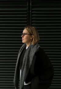 A person wearing glasses, a grey scarf, and a black coat stands against a dark slatted background. Their hair is shoulder-length and straight.
