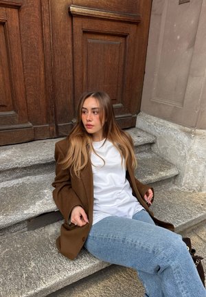 Joven mujer con el pelo largo, blazer marrón, camisa blanca y vaqueros azules sentada en escalones de piedra junto a una gran puerta de madera, mirando hacia un lado.