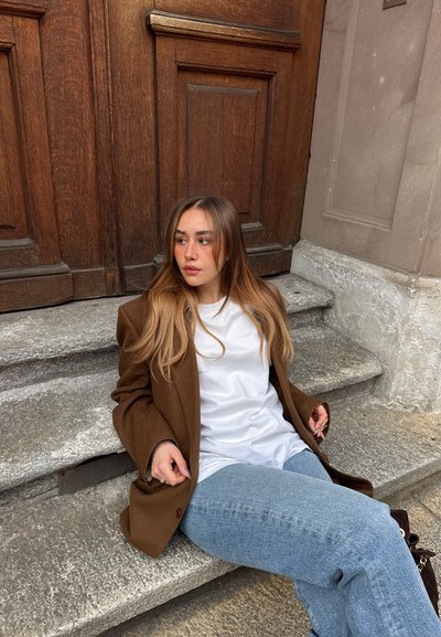 Joven mujer con el pelo largo, blazer marrón, camisa blanca y vaqueros azules sentada en escalones de piedra junto a una gran puerta de madera, mirando hacia un lado.