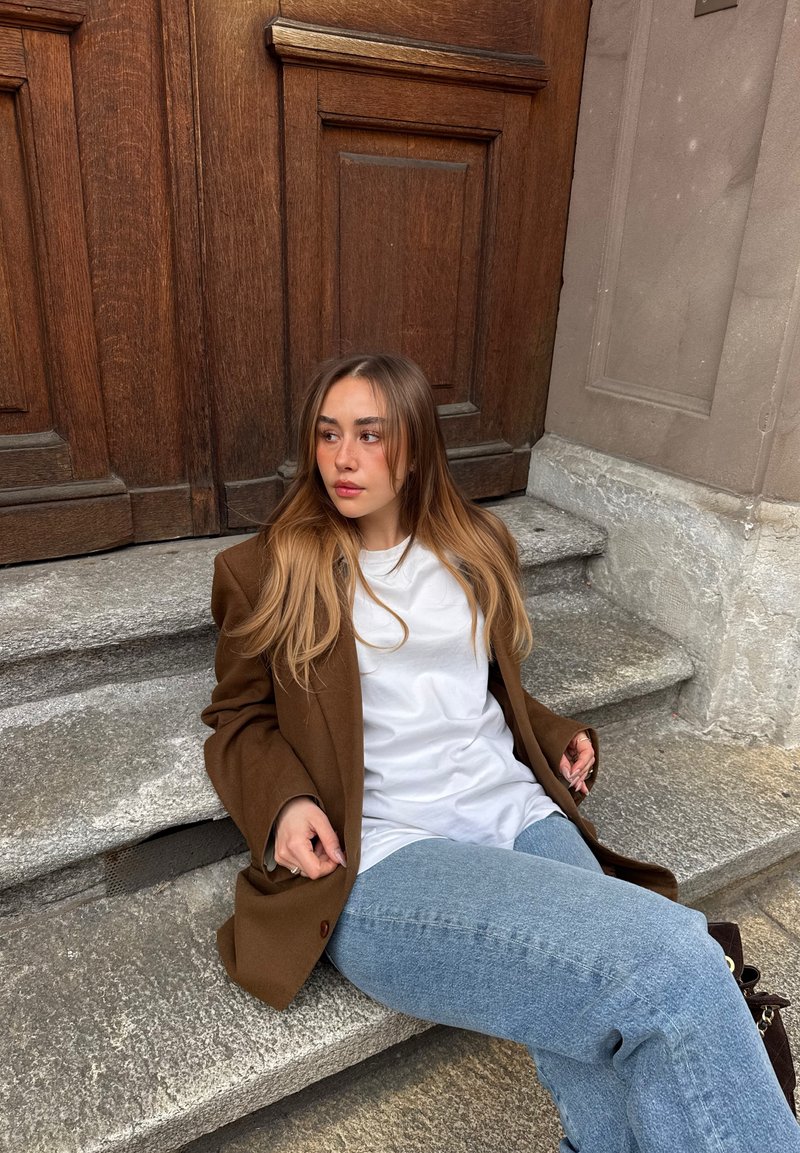 Jeune femme aux cheveux longs portant un blazer marron, une chemise blanche et un jean bleu assise sur des marches en pierre près d'une grande porte en bois, regardant sur le côté.
