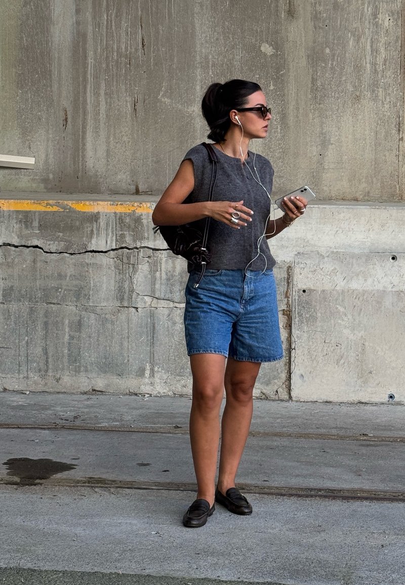 Top grigio senza maniche, pantaloni shorts in denim blu e mocassini neri. Tiene uno smartphone, indossa auricolari, con una borsa scura a tracolla.