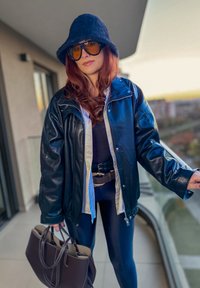 Chaqueta de cuero negro con acabado brillante, combinada con una camisa clara. Sombrero tipo pescador de fieltro azul oscuro, gafas de sol grandes y bolso marrón.