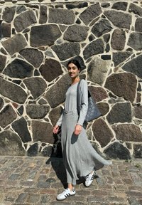 Robe grise à manches longues avec une jupe fluide et des détails de ceinture, associée à des sneakers blanches et un sac à bandoulière tissé gris, devant un mur en pierre.