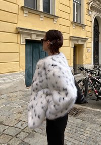 Bela umetna kožuh z črnimi lisami, veliko zasnovo in puhasto teksturo. Oblečena s črnimi hlačami; utišana urbana ozadje s kolesi.