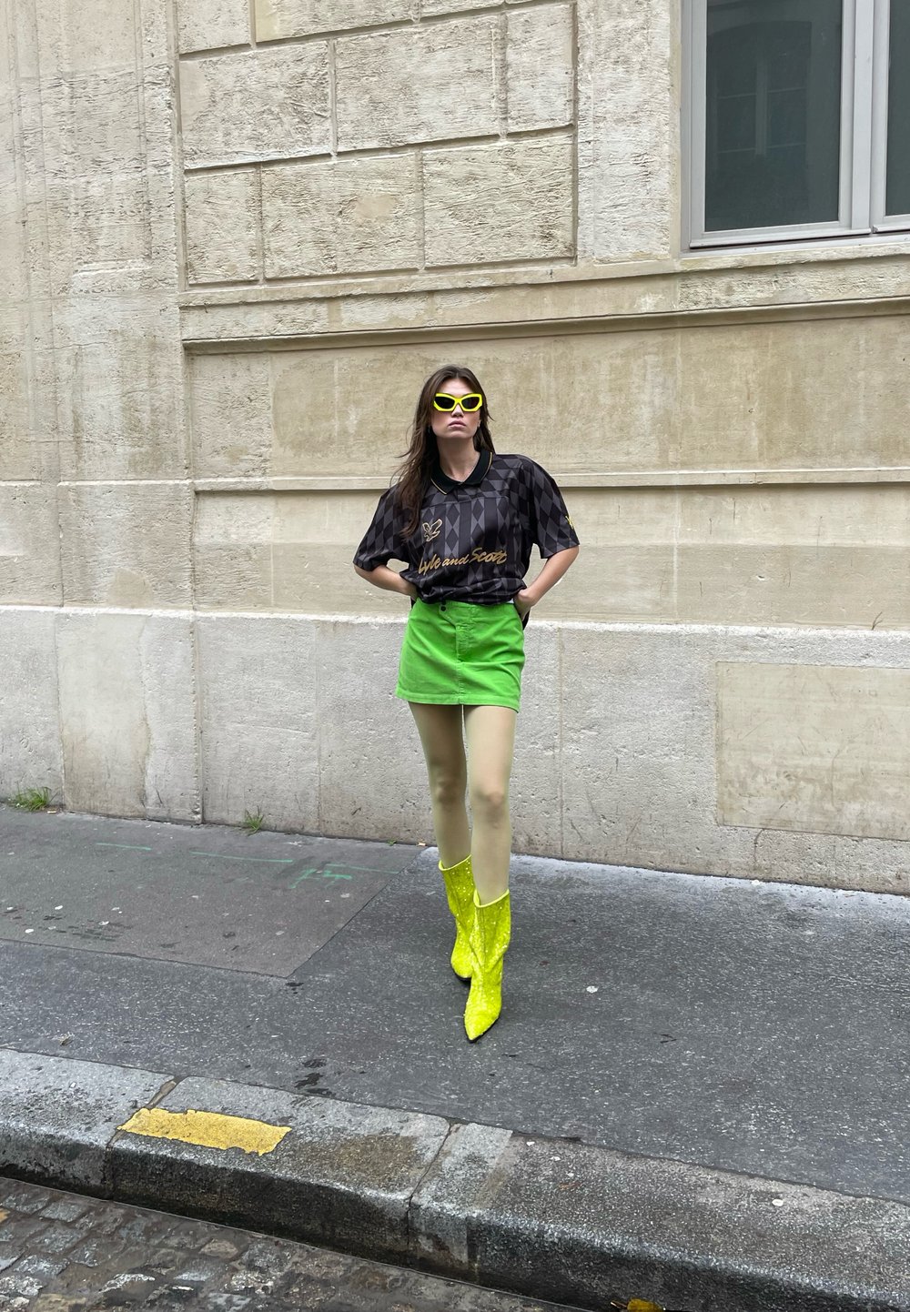 Schwarzes kurzärmliges Oberteil mit einem Diamantmuster und goldenem Text, grüner Minirock, transparente hellgrüne Strumpfhosen und glänzende neongelbe Stiefel bis zum Knie.