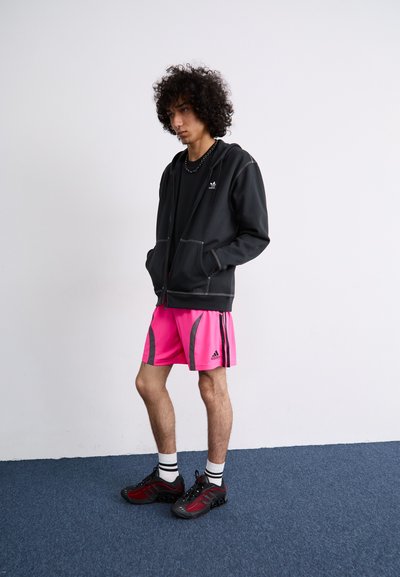 Sudadera negra con cremallera y costuras grises, combinada con shorts deportivos rosa brillante con detalles negros. Zapatillas deportivas negras y rojas.