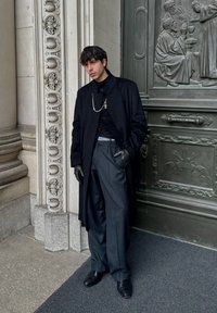 Man in een zwarte jas, lederen handschoenen en grijze broek staat bij een sierlijke stenen zuil en een gedetailleerde metalen deur met religieuze reliëfs.