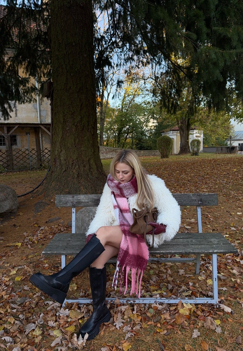 Hvid pels, lyserødt ternet tørklæde, sorte knælange støvler, sidder på en træbænk omgivet af efterårsblade og træer. Brun taske ved siden af.