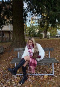 Hvid pels, lyserødt ternet tørklæde, sorte knælange støvler, sidder på en træbænk omgivet af efterårsblade og træer. Brun taske ved siden af.