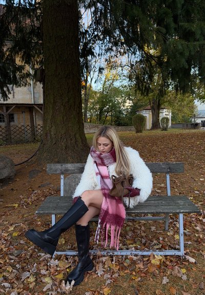 Abrigo blanco y esponjoso, bufanda rosa con cuadros, botas negras hasta la rodilla, sentada en un banco de madera entre hojas y árboles otoñales. Bolso marrón al lado.