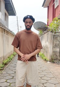 T-shirt en coton marron à manches courtes, associé à un short beige. La personne porte un béret noir avec des lettres blanches et des bagues argentées.