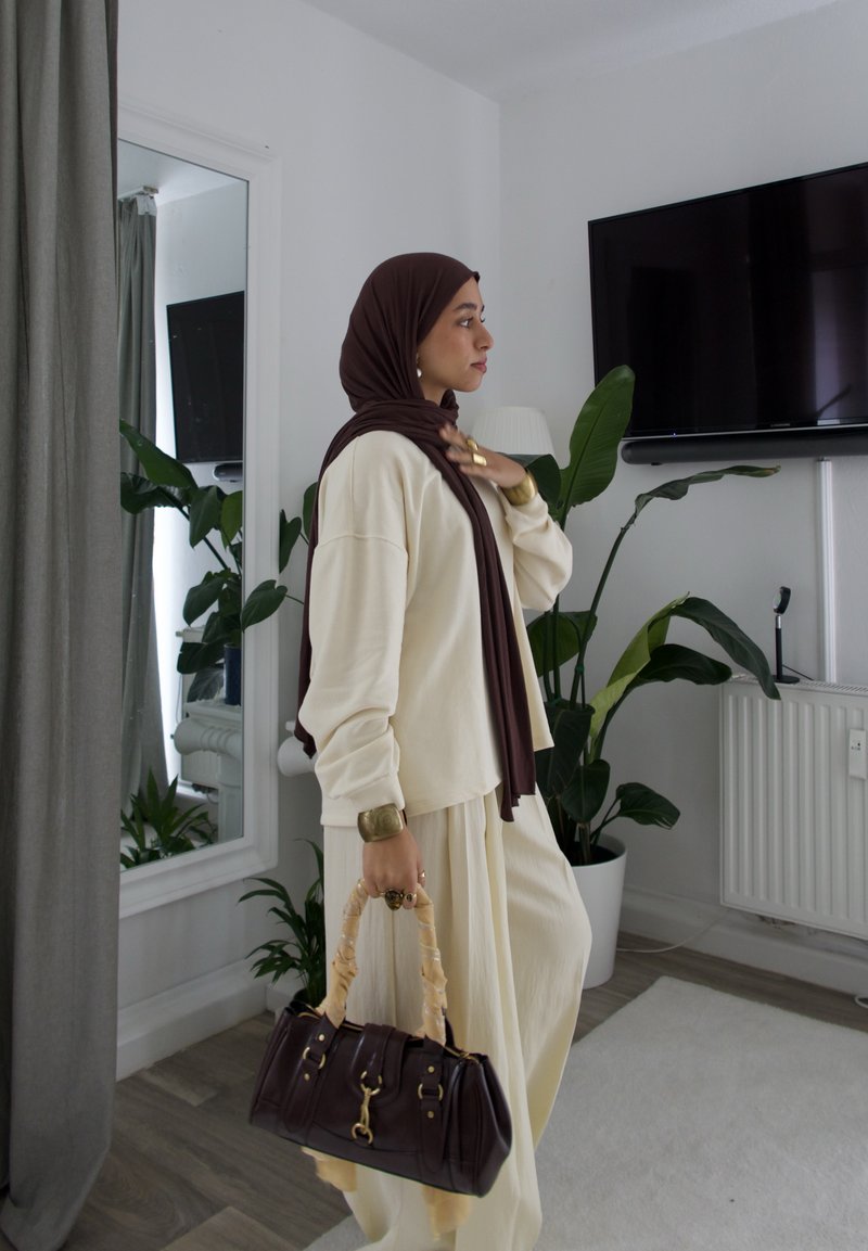 Maglietta oversize color crema e pantaloni abbinati, accessorati con una borsa marrone scuro e gioielli dorati, sullo sfondo di piante d'appartamento.