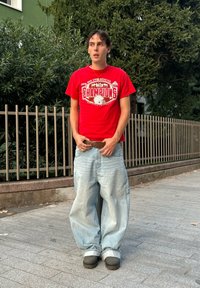 T-shirt rouge avec un motif "Champions", jean baggy en denim bleu clair et chaussures de type slip-on foncées. Debout en extérieur avec un fond de feuillage vert.