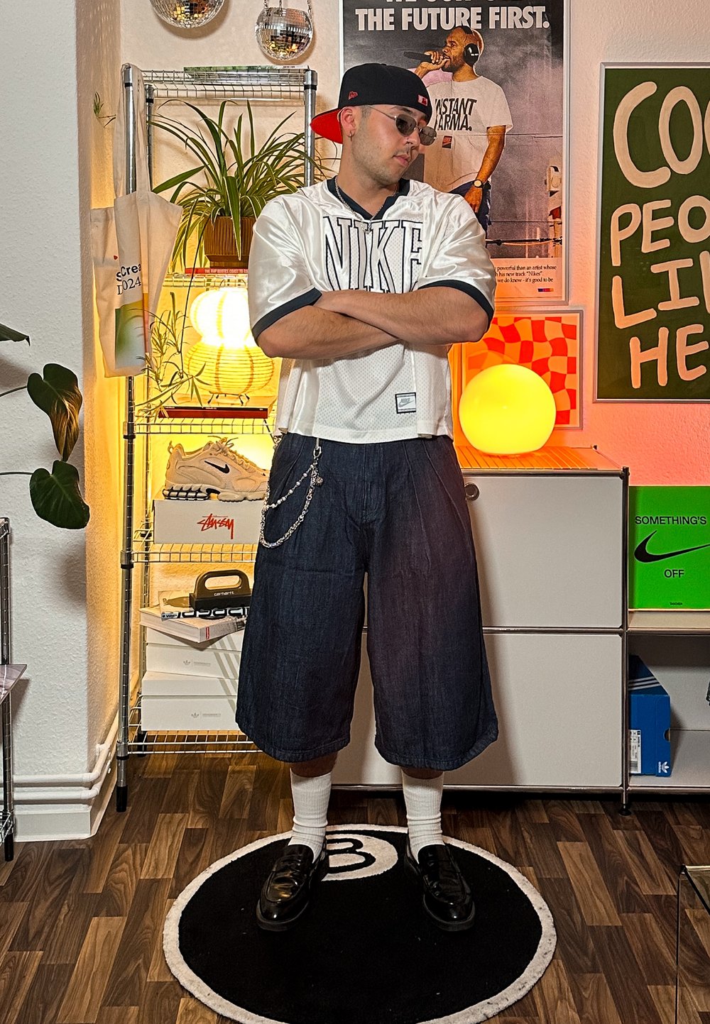 Stříbrný oversized Nike dres, tmavé denimové kraťasy, řetězový detail, černé mokasíny a bílé crew ponožky. V pozadí jsou světla a dekorace.