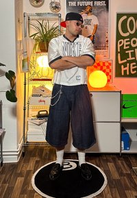 Stříbrný oversized Nike dres, tmavé denimové kraťasy, řetězový detail, černé mokasíny a bílé crew ponožky. V pozadí jsou světla a dekorace.